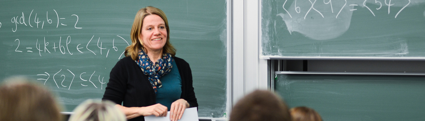 Lecturer stood smiling holding paper in front of green chalkboard