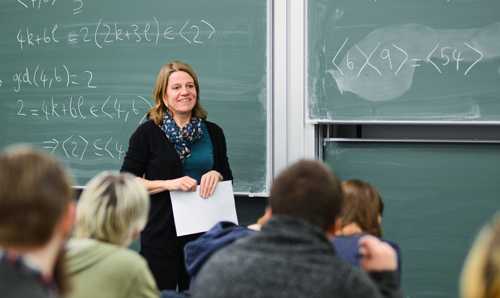 Lecturer stood smiling holding paper in front of green chalkboard